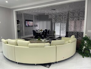 A modern living room with a curved white sofa, a glass coffee table, and a dining area with a black table and chairs. - Beautiful villa for big family  (Courtice)