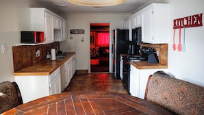 Cocina compacta con gabinetes blancos, microondas y refrigerador. Un comedor con sillas está en primer plano.