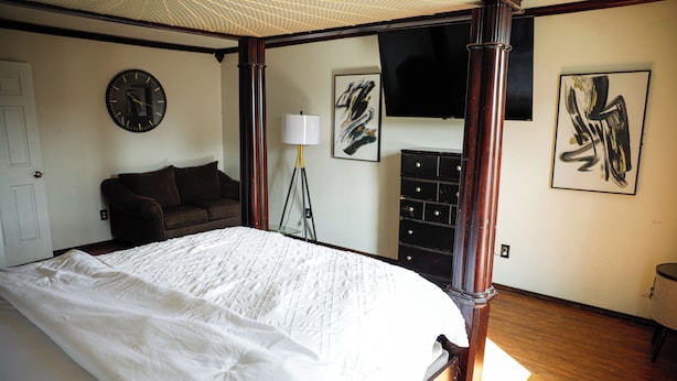 Un dormitorio con una cama grande, piso de madera, un reloj, una lámpara y obras de arte en las paredes.