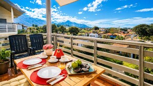 Une table à manger installée à l’extérieur, avec des chaises, des assiettes, des tasses et une théière rouge, d’où l’on peut admirer un paysage montagneux.