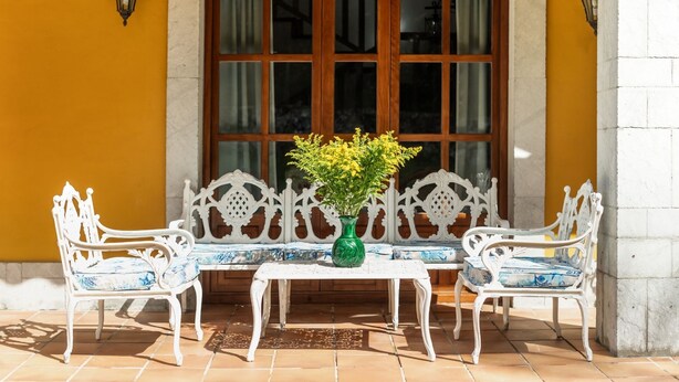 Eine Terrasse mit weißem Tisch und Stühlen, einer grünen Vase mit gelben Blumen und einem Fenster mit hölzernen Rollläden.