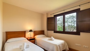 Une chambre d’hôtel avec deux lits simples, une table de chevet avec une lampe, une fenêtre avec des volets et une vue sur les arbres à l’extérieur.