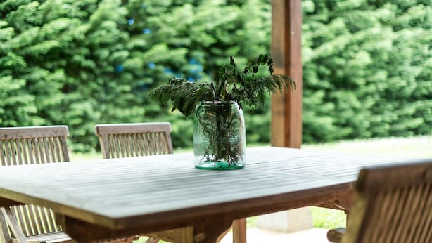 Une table en bois sur laquelle est posé un vase en verre contenant des plantes vertes, le tout installé à l’extérieur, avec un banc en bois et un décor de feuillage vert et luxuriant en arrière-plan.