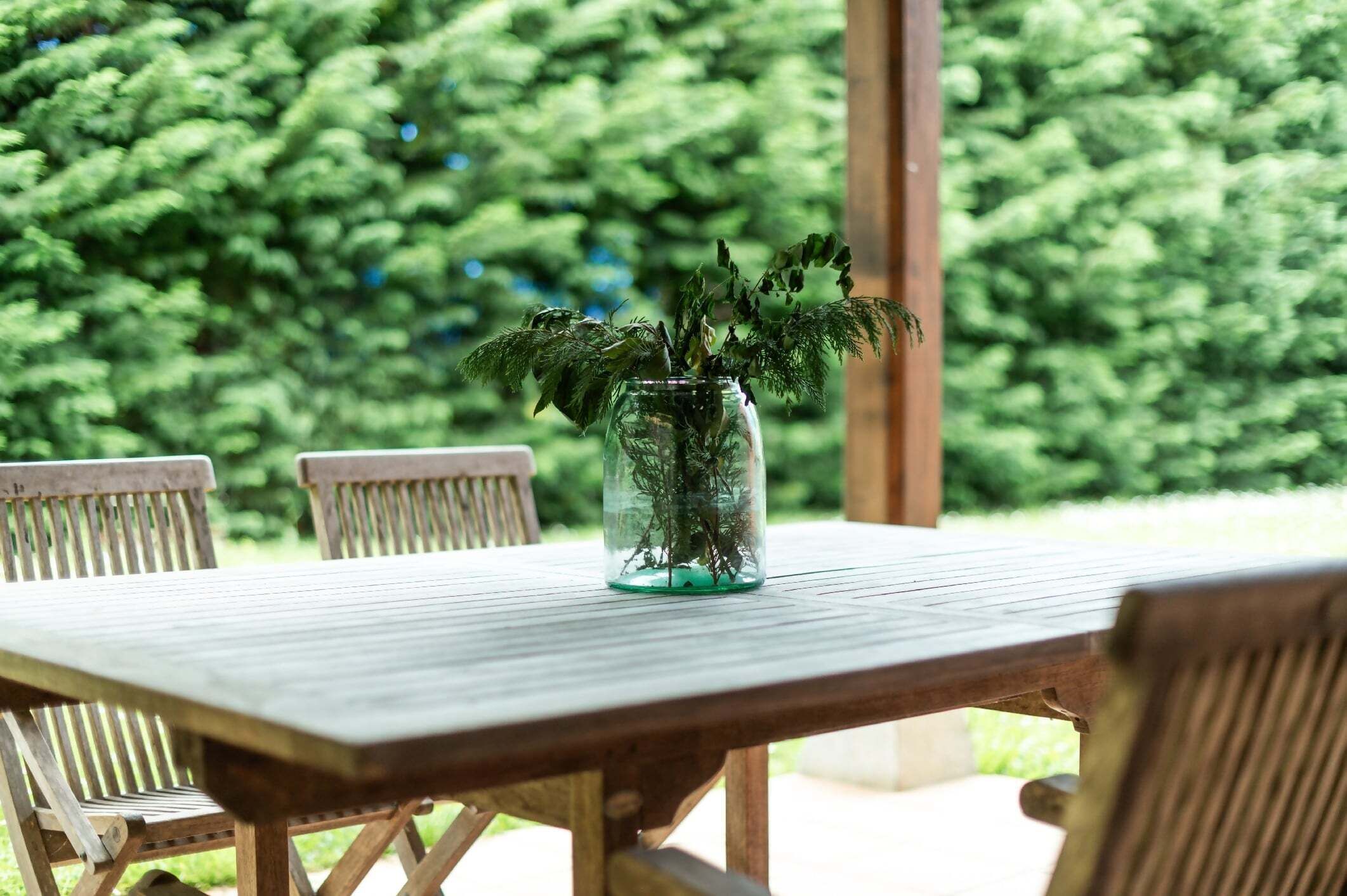 A wooden table with a glass vase containing green plants, surrounded by wooden chairs.