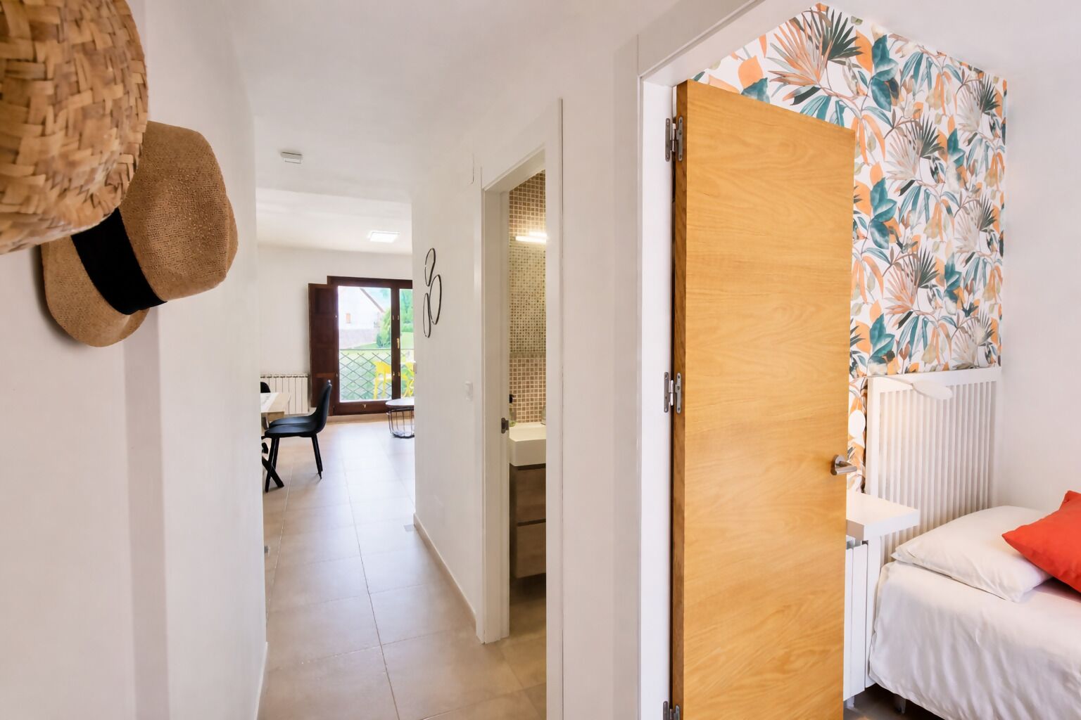 Un couloir mène à une chambre dont la porte est en bois, ainsi qu’à un coin repas avec une table et des chaises, et offre une vue sur un balcon.