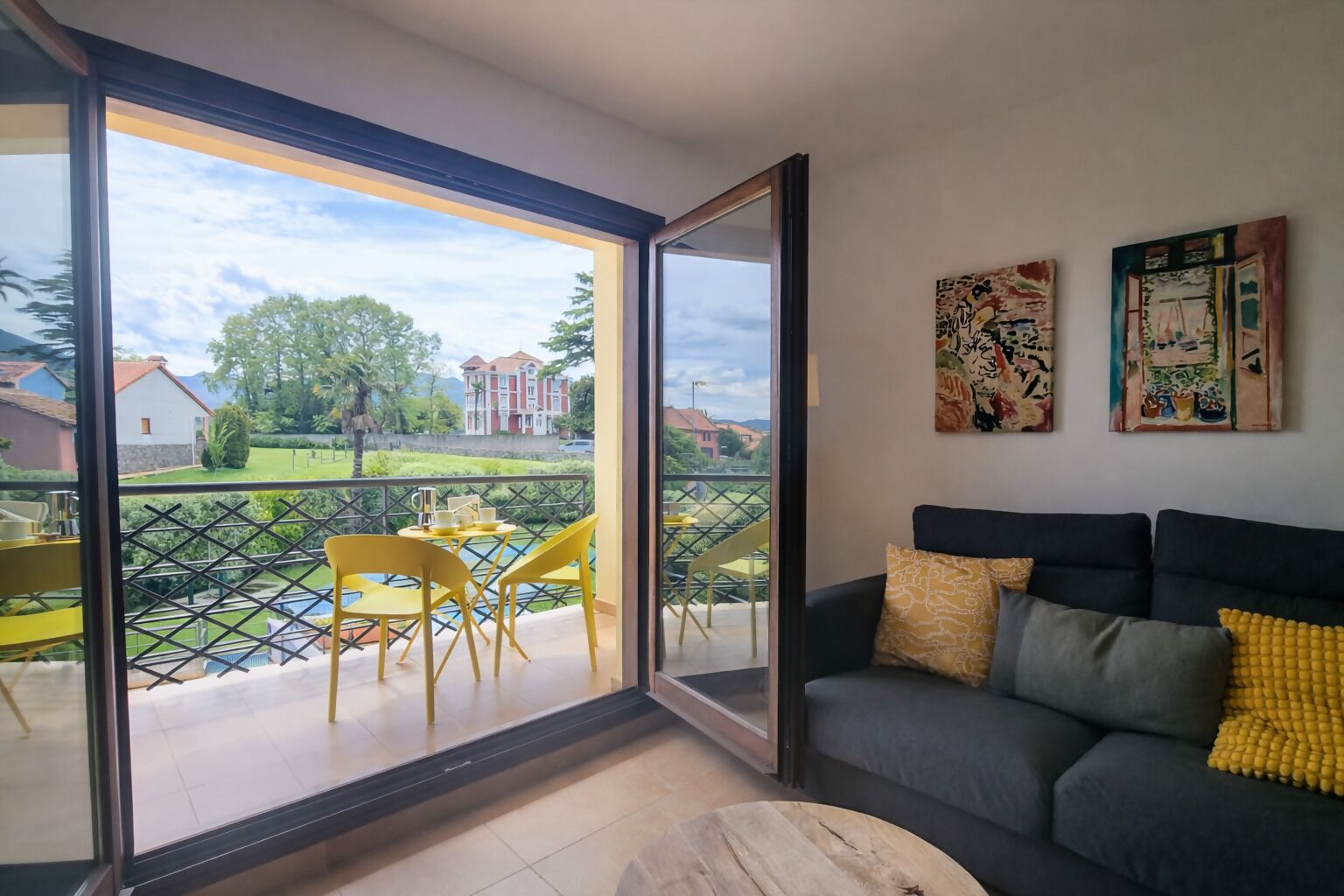 Una sala de estar moderna con un ventanal corredizo que da a un balcón. La habitación cuenta con un sofá gris oscuro, cojines amarillos y grises, una mesa de centro de madera y dos cuadros abstractos en la pared.