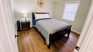 Un dormitorio con cama, mesita de noche, lámpara, ventilador de techo, decoración de pared con brújula y ventana con persianas.