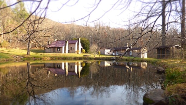 Un chalet au bord du lac, dont l’image se reflète dans l’eau, entouré d’arbres et d’une zone herbeuse.