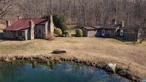 Une cabane rustique avec un toit en métal, une cheminée en pierre et un petit étang au premier plan.
