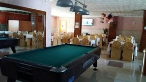 Una mesa de billar en una sala con mesas dispuestas para comer, sillas con fundas blancas y un televisor en la pared.