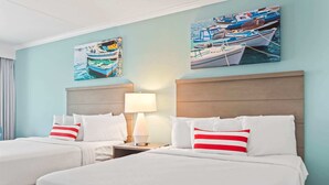 Quarto de hotel com duas camas, uma mesa de cabeceira com abajur e duas pinturas de barcos na parede.