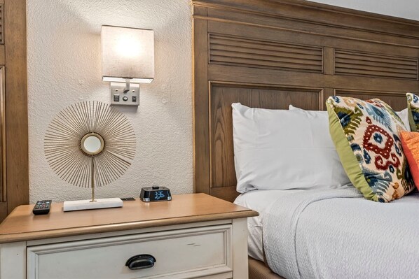 A neatly made bed with white and colorful pillows, a bedside table with a clock, a remote control, and a wall-mounted light fixture.