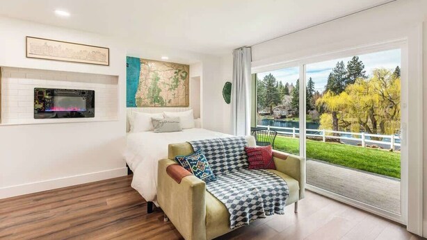 A modern bedroom with a bed, a sofa, a map on the wall, and a view of a park through large windows. - Riverfront Studio - Private Patio & Pet Friendly! (Bend)