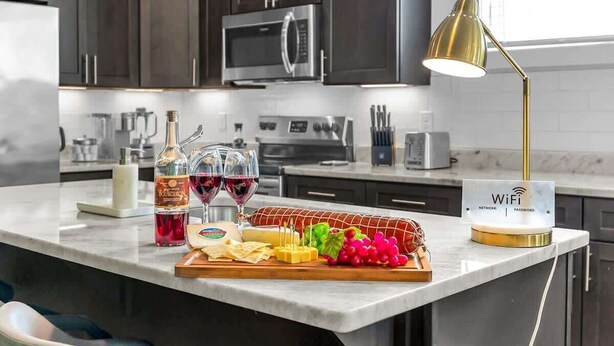 Een keuken met donkere kastjes, roestvrijstalen apparatuur, een marmeren aanrecht, een bord met het WiFi-wachtwoord, en diverse soorten eten en drinken.
