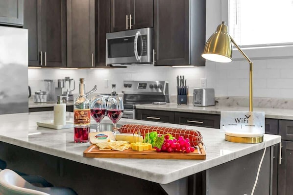 A kitchen with dark cabinets, stainless steel appliances, a marble countertop, a WiFi password sign, and a variety of food and drink items.