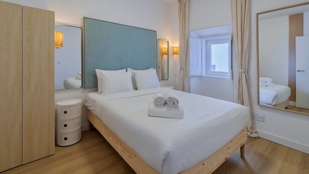 Ein modernes Hotelzimmer mit einem Bettgestell aus Holz, weißer Bettwäsche, einem großen Spiegel und einem Fenster mit Vorhängen.