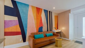 Ein modernes Wohnzimmer mit einem farbenfrohen abstrakten Wandbild, einer braunen Ledersofa mit türkisfarbenen Kissen, einem kleinen Holztisch und einem Schreibtisch mit Lampe.