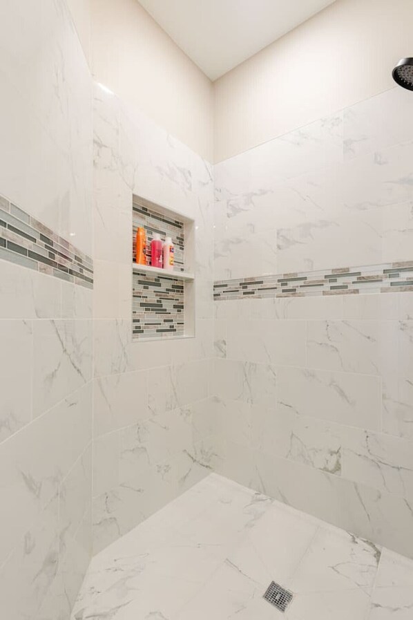 A modern bathroom with white marble walls and a built-in shelf with toiletries.