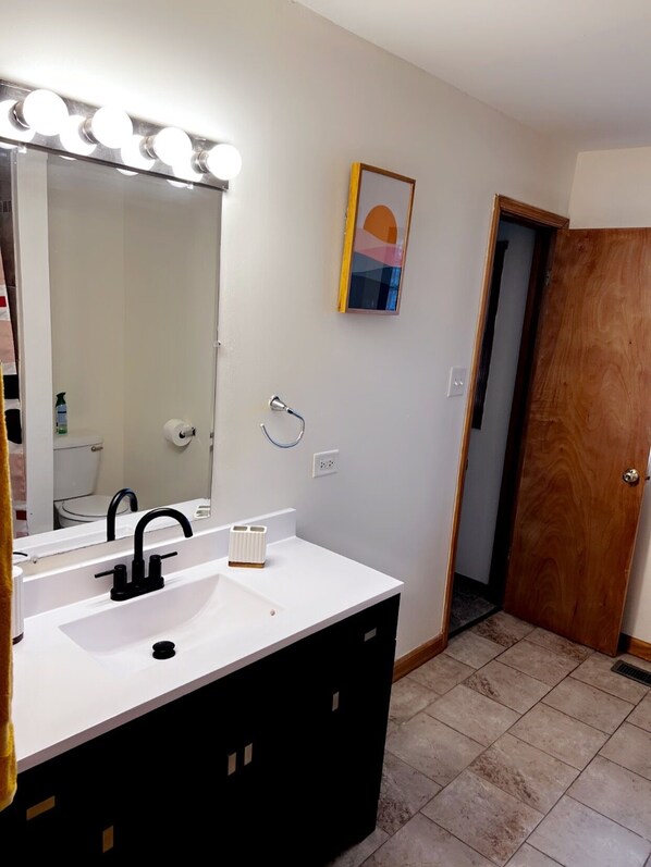 Un baño con lavamanos blanco, mueble negro y espejo con luces. Se observa una puerta y un pequeño cuadro enmarcado en la pared.