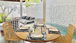A dining area with a round table set for four, surrounded by wicker chairs, with a stone wall and a pool in the background. - CALM VILLA SEMINYAK (Seminyak)