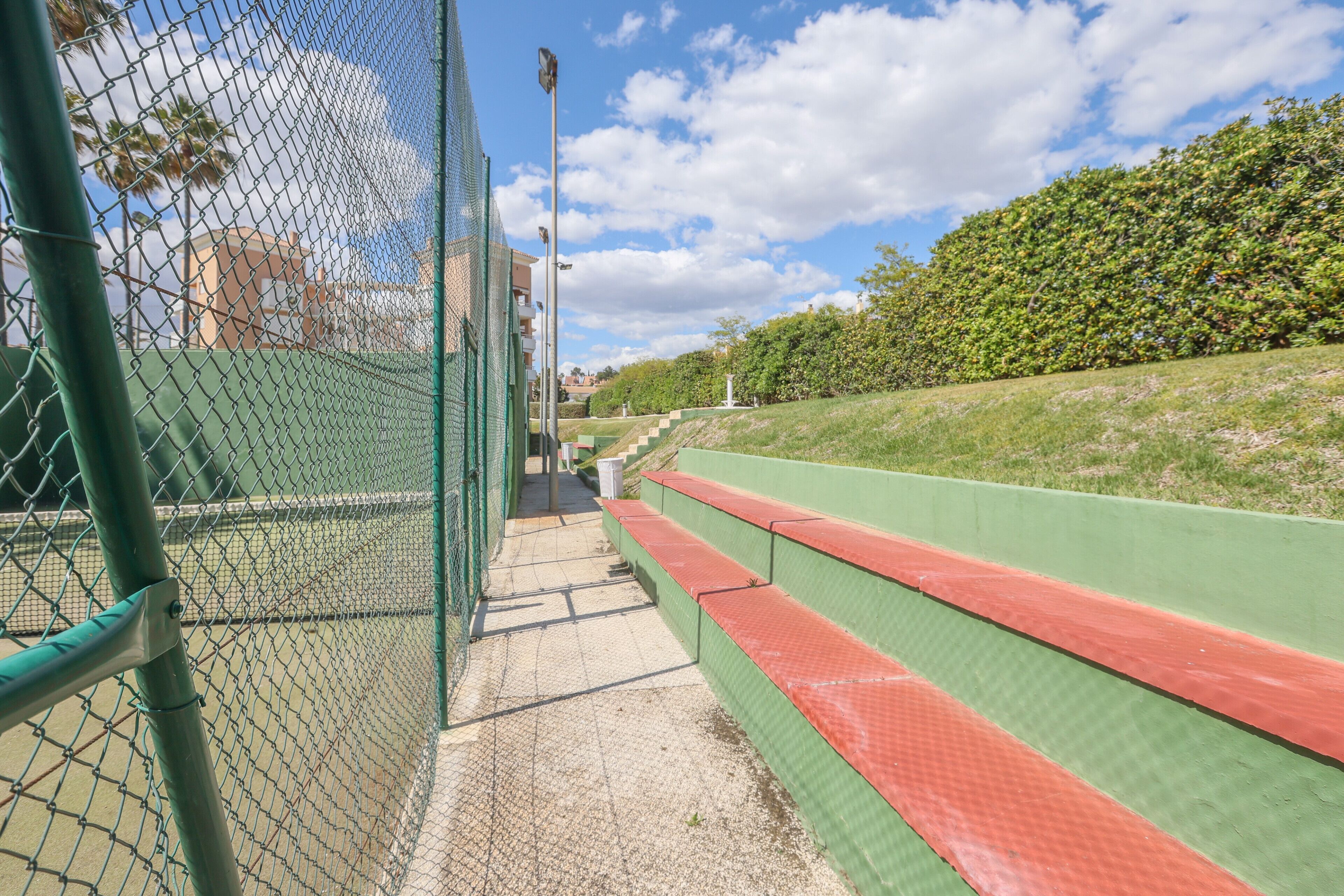 Een tennisbaan met een hekken, een zitgedeelte in groen en rood, en een heldere lucht.