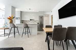 A modern kitchen with a central island, bar stools, a dining table with chairs, and a flat-screen TV mounted on the wall.