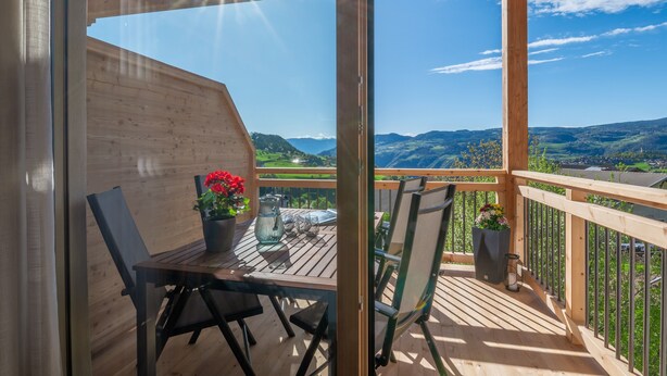 Um deck de madeira com mesa e cadeiras, plantas em vasos e vista para colinas ao longe.