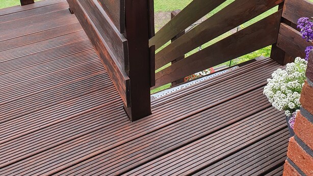 Um deck de madeira com guarda-corpo, com vista para um jardim ajardinado, uma estrada e vegetação.