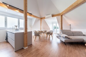 A modern kitchen with a central island, a dining area with a round table and chairs, a sofa, and large windows.