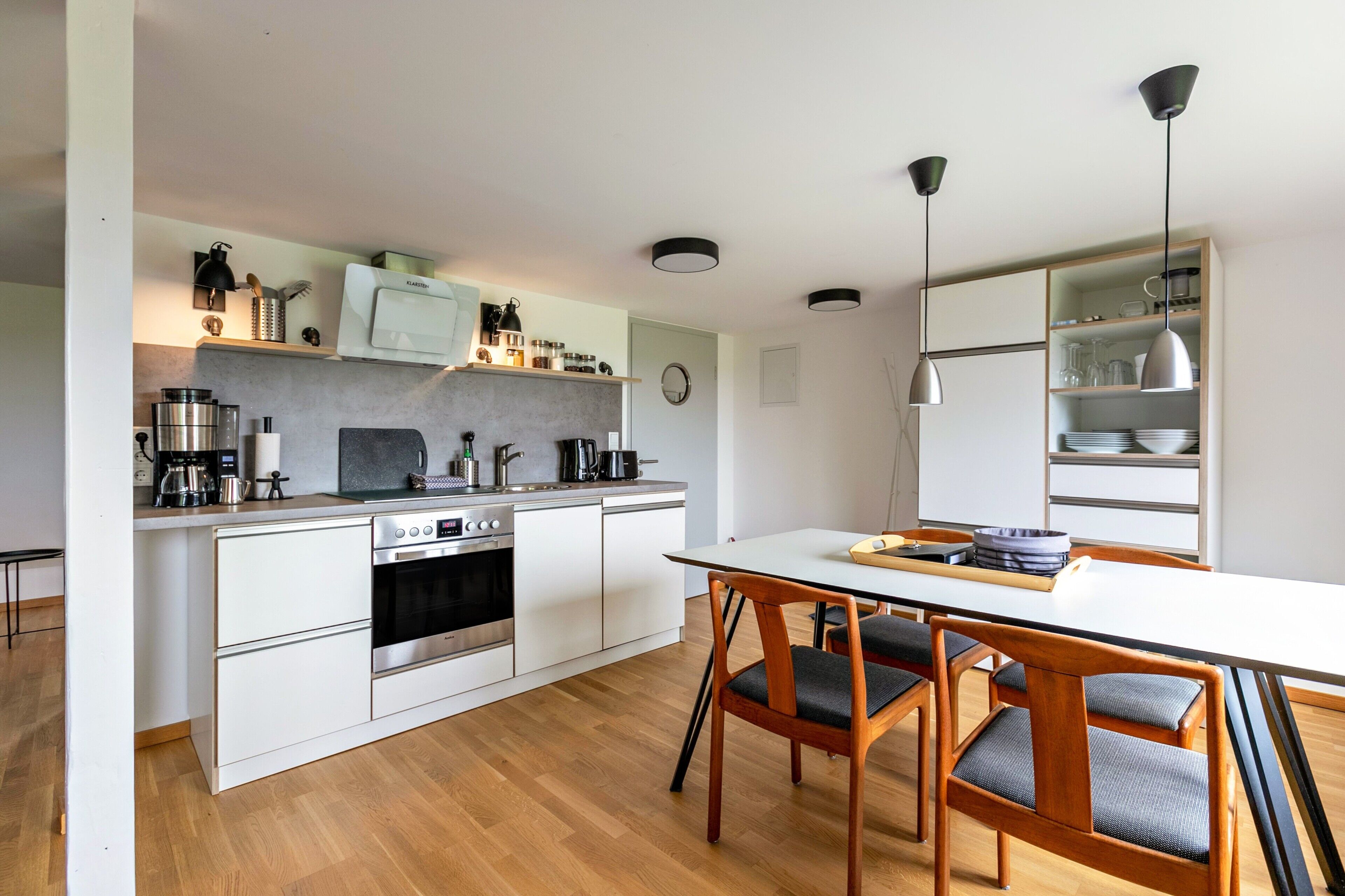 Una cocina moderna con armarios blancos, horno integrado y una zona de comedor con mesa blanca y sillas de madera.