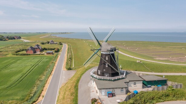 A windmill with a road and fields in the foreground, and a body of water in the background.