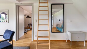 A room with a wooden ladder, a mirror, a blue chair, a white cabinet, and a door leading to another room.