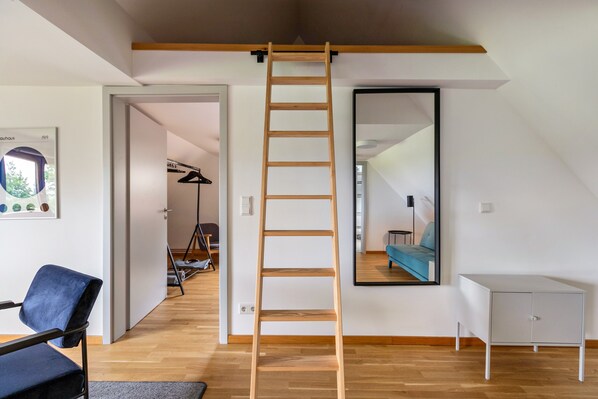 A room with a wooden ladder, a mirror, a blue chair, a white cabinet, and a door leading to another room.
