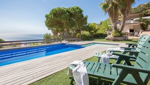 A swimming pool with a wooden deck, green lounge chairs, and a view of the sea.