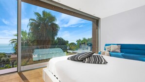 A bedroom with a large sliding glass door leading to a balcony with a view of the ocean and palm trees.