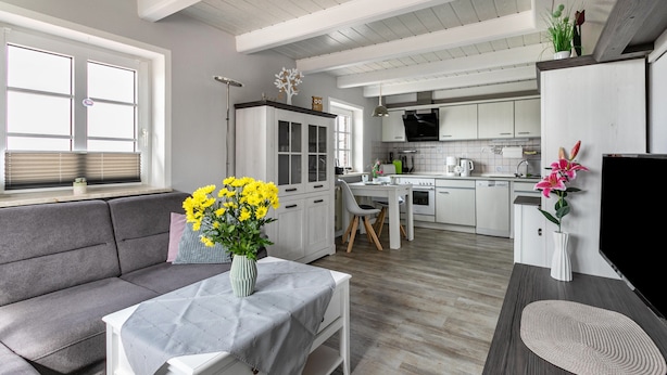Una cucina moderna con mobili bianchi, una zona pranzo con un divano grigio e un tavolino con un vaso di fiori gialli.