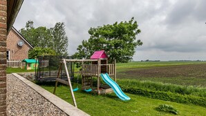 Un’area giochi in legno con scivolo, altalene e recinzione, adiacente a un edificio in mattoni e a un campo.
