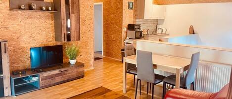 A cozy living room with a TV, wooden furniture, a red sofa, and a kitchen area with a dining table and chairs.
