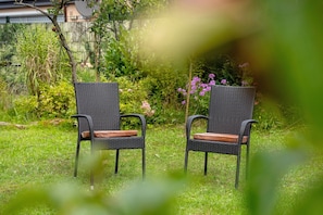 Two wicker chairs with wooden seats in a grassy outdoor area with a garden and a fence in the background.