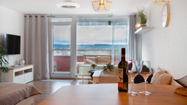 Un salon moderne avec une table en bois, deux verres à vin, une bouteille de vin, un téléviseur et un balcon avec vue.
