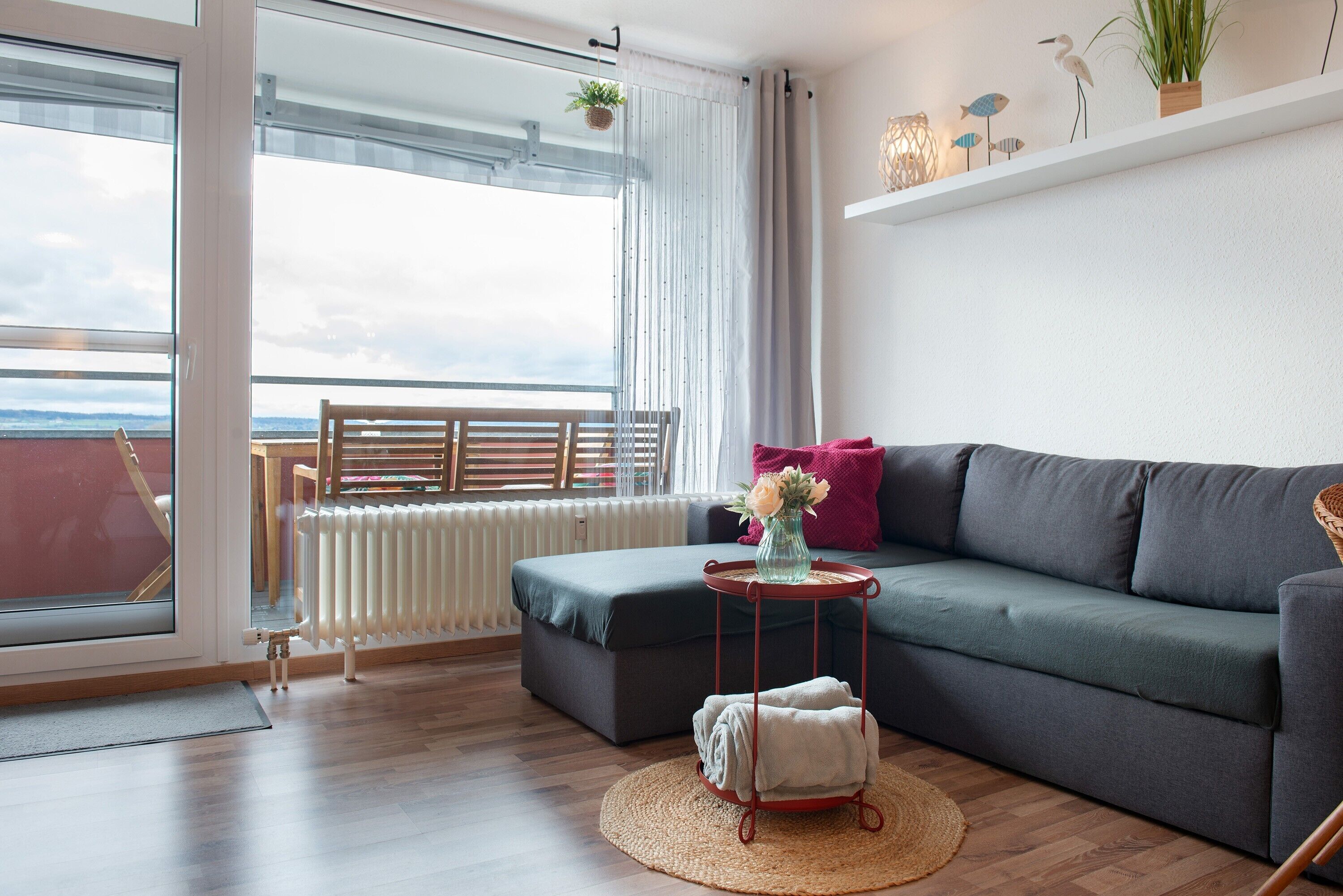 A living room with a sofa, a small side table, a radiator, a window with curtains, and a view of the sea.