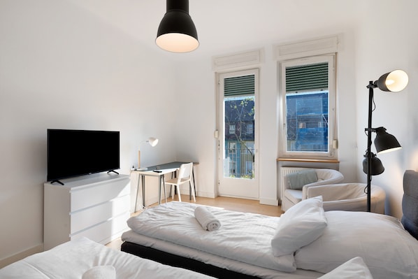 A bedroom with a bed, a desk, a chair, a TV, and two windows.