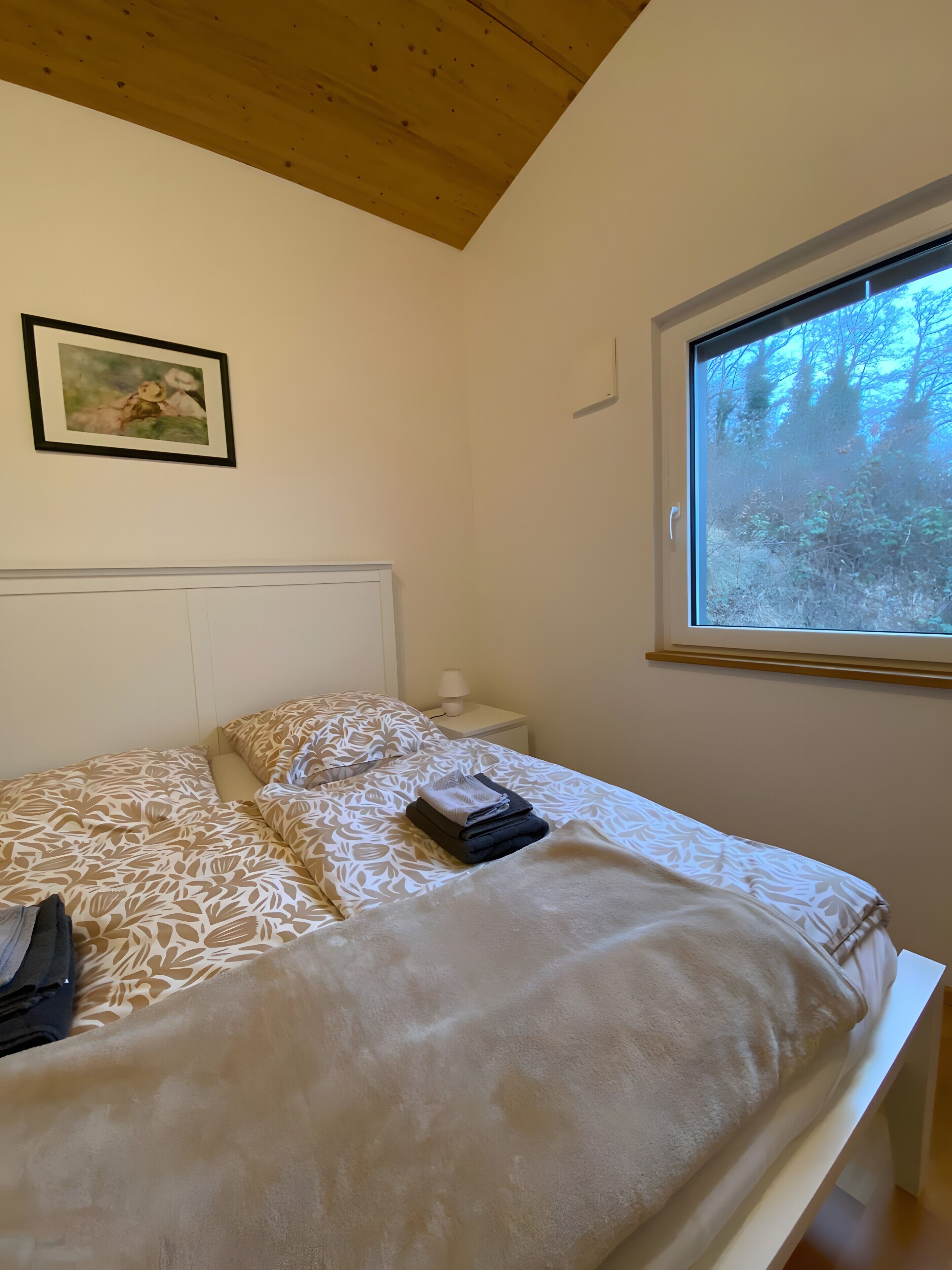 Ein Schlafzimmer mit Bett, Nachttisch, Lampe, gerahmtem Bild an der Wand und Fenster mit Blick auf Bäume.