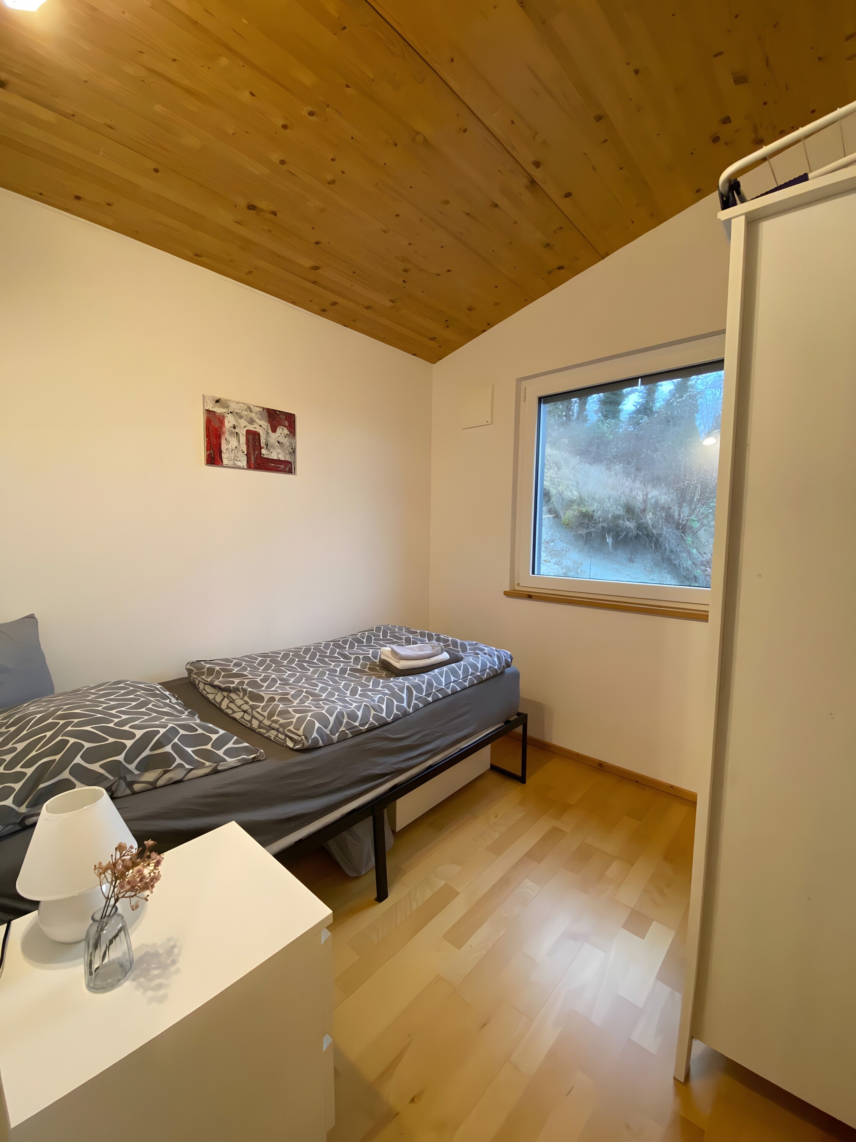 Ein kleines Zimmer mit Bett, Fenster, weißer Wand und Holzboden.