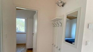 Een gang met houten vloer, witte wanden, een deur die naar een andere kamer leidt, en een spiegel aan de muur.