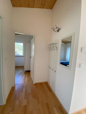 Un pasillo con suelo de madera, paredes blancas, una puerta que da a otra habitación y un espejo en la pared.