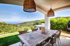 A wooden table with wicker chairs and hanging wicker lamps, set on a terrace with a view of the sea and hills.