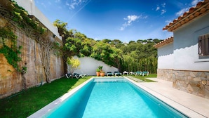Une piscine rectangulaire avec une terrasse en bois au premier plan, entourée d’un mur blanc recouvert de plantes grimpantes, d’un mur en pierre et d’un petit bâtiment doté d’un toit en tuiles.