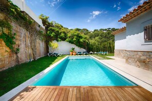 A rectangular swimming pool with a wooden deck in the foreground, surrounded by a white wall with climbing plants, a stone wall, and a small building with a tiled roof. - Villa 'Casa Chula' with Sea View, Wi-Fi and Air Conditioning (Sa Riera)