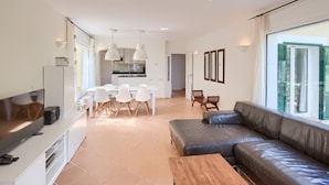 Un salon moderne avec un canapé en cuir, un coin repas avec des chaises blanches et une cuisine en arrière-plan.